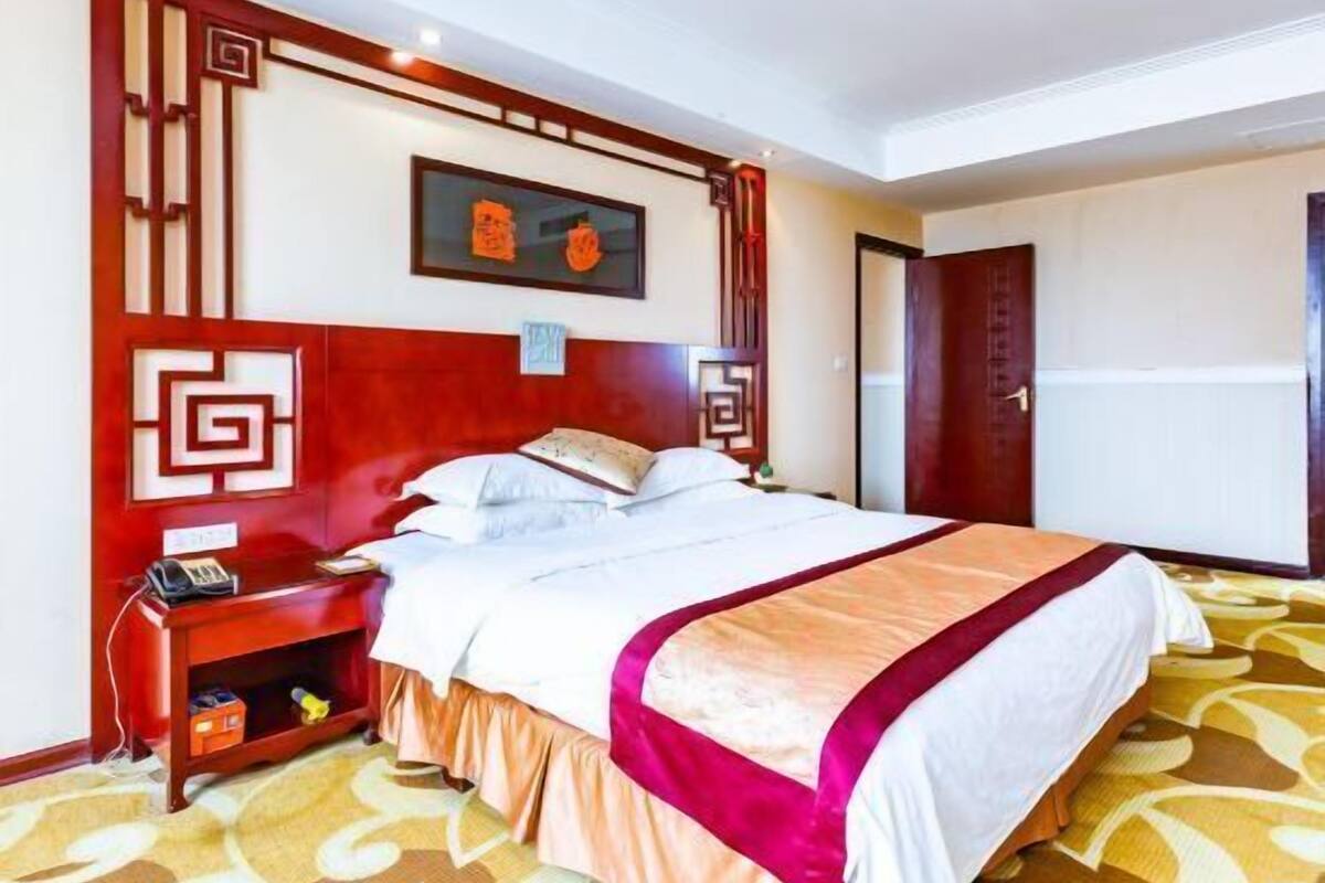 Habitación de hotel con una cama grande, cabecera de madera, mesita de noche y un panel decorativo rojo en la pared.