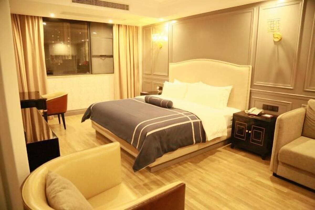 A hotel room with a large bed, a bedside table with a lamp, a chair, and a view of the city through the window.