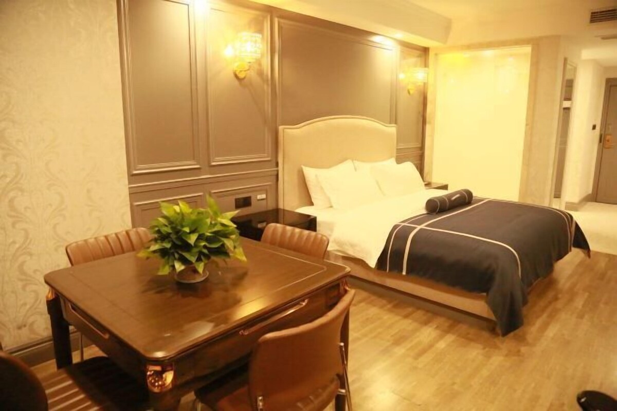 A hotel room with a wooden dining table, a bed with a black and white bedspread, and a potted plant on the table.