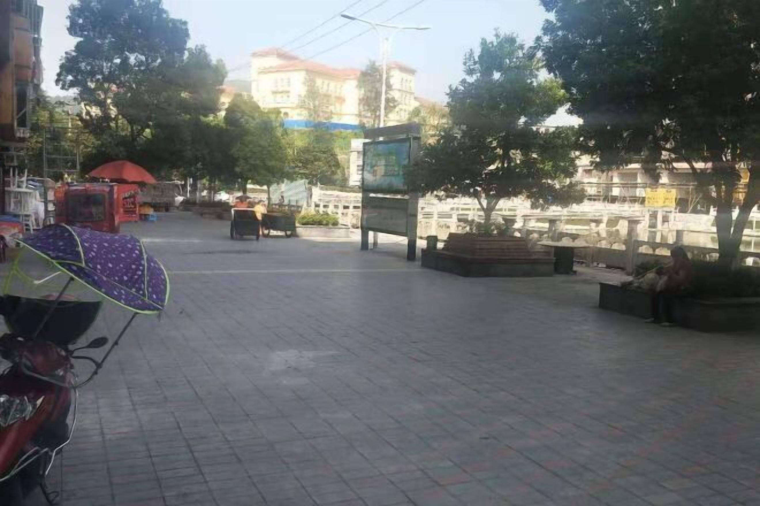 Une place dallée, agrémentée de bancs, d’arbres et d’un scooter garé.