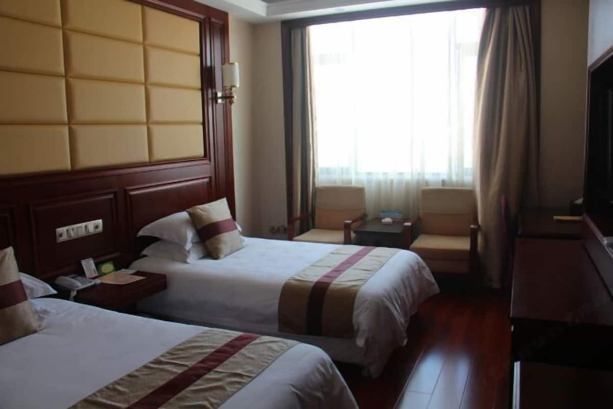 A hotel room with two beds, a wooden headboard, a small table, a chair, and a large window with curtains.