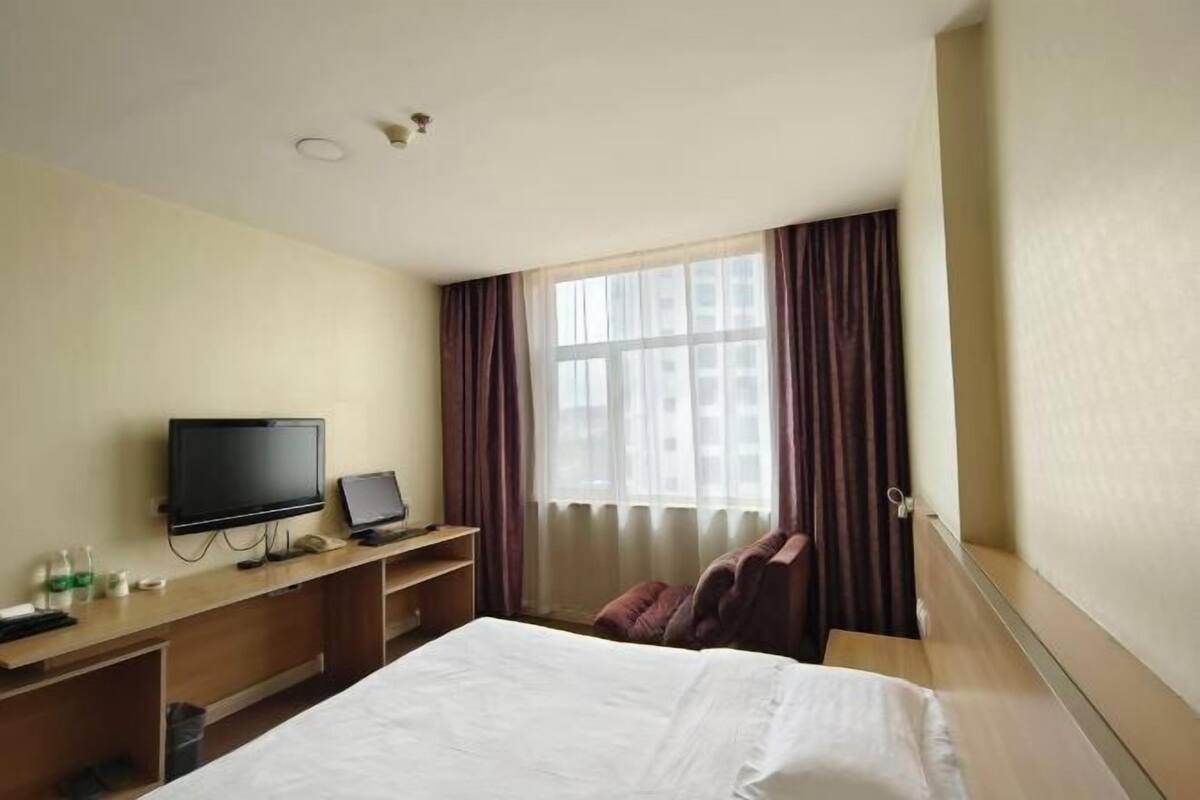 Ein Hotelzimmer mit einem Bett, einem Schreibtisch mit Fernseher und Laptop, einem Fenster mit Vorhängen und einem Sessel.