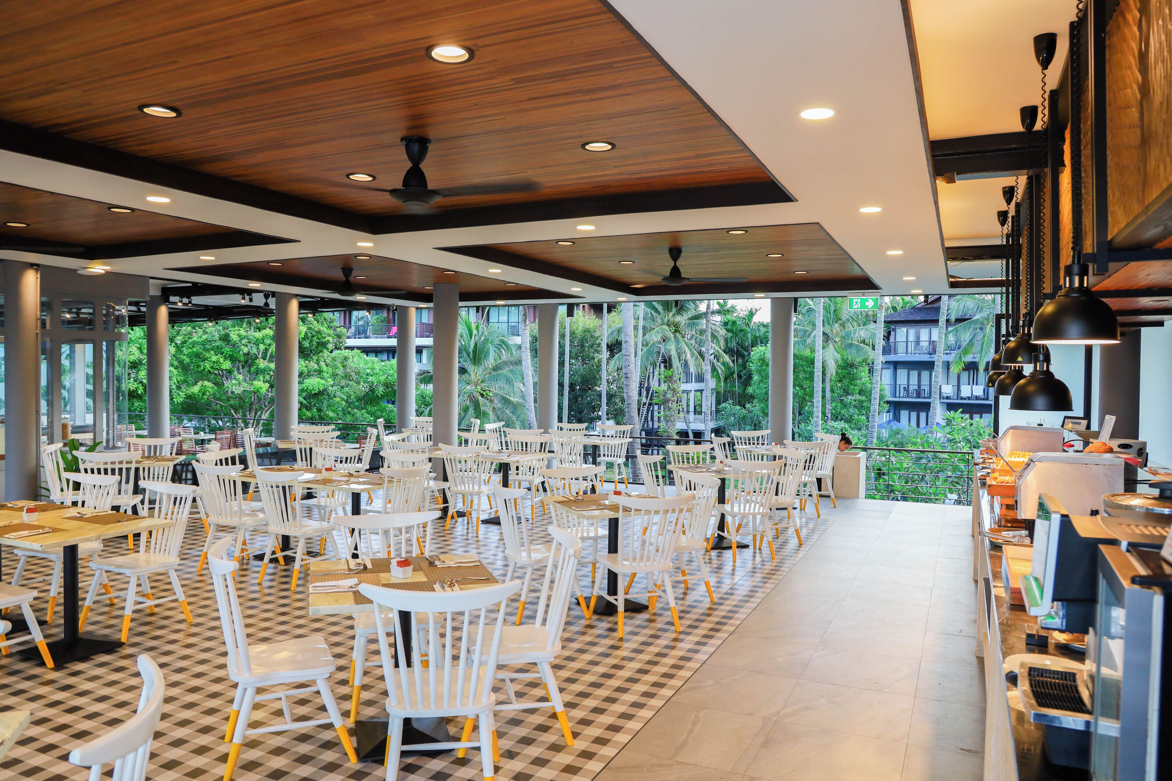 A spacious indoor dining area with white tables and chairs, large windows offering views of greenery, and ceiling fans.