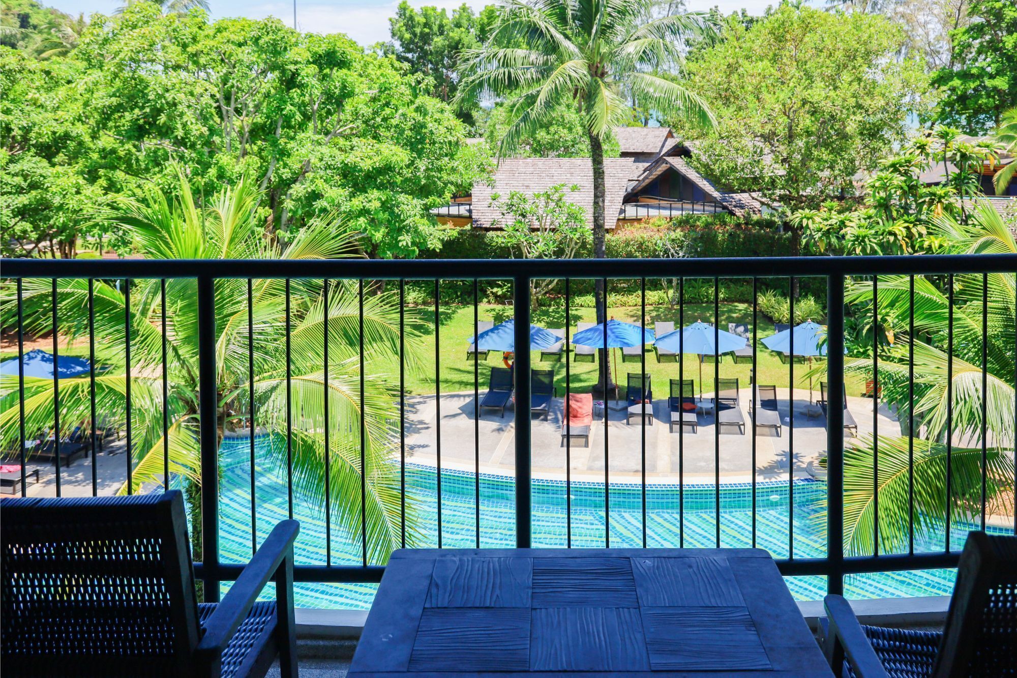 A poolside view with lounge chairs and umbrellas, surrounded by lush greenery and a building in the background.