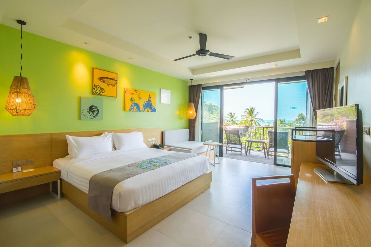 A hotel room with a large bed, a desk, a TV, and a balcony with a view of palm trees.
