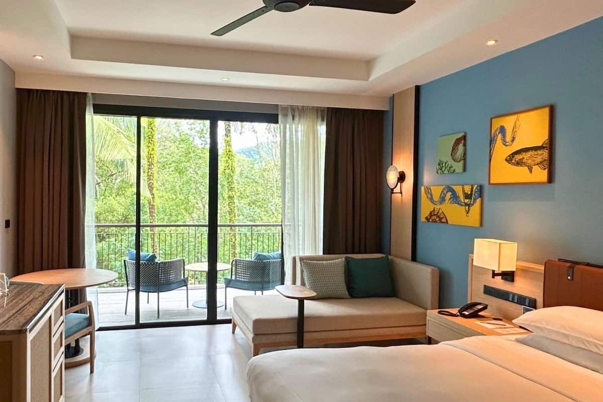 A hotel room with a bed, a desk, a chair, a small table, and a balcony with chairs.