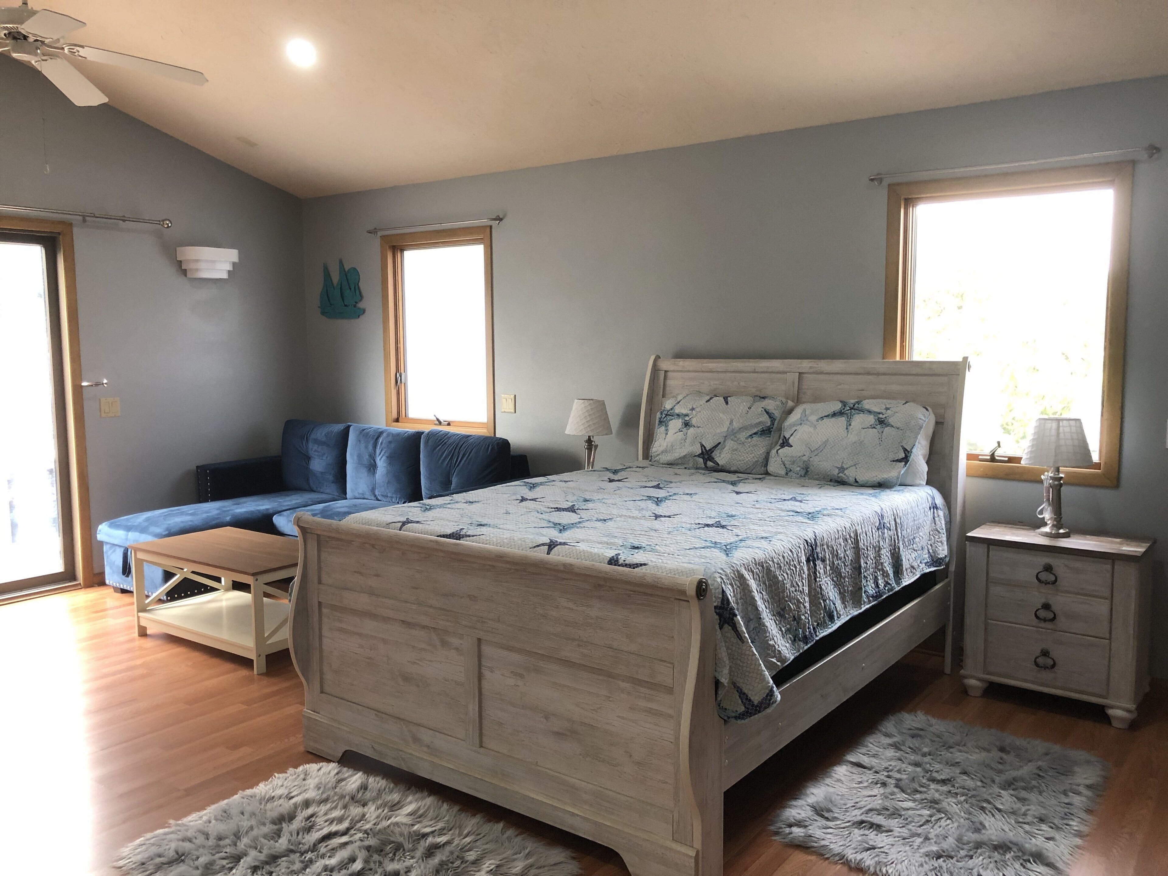 A bedroom with a wooden bed, a blue sofa, a small table, and a nightstand. There are two windows providing natural light.