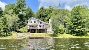 A two-story house with a dock extending into a lake, surrounded by dense trees. - Lakefront Escape - Private Dock - Pet Friendly (Albrightsville)