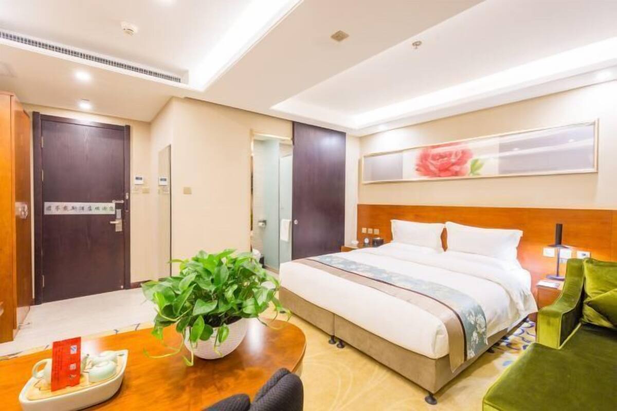 A hotel room with a bed, a desk with a plant, a green armchair, and a wall-mounted artwork.