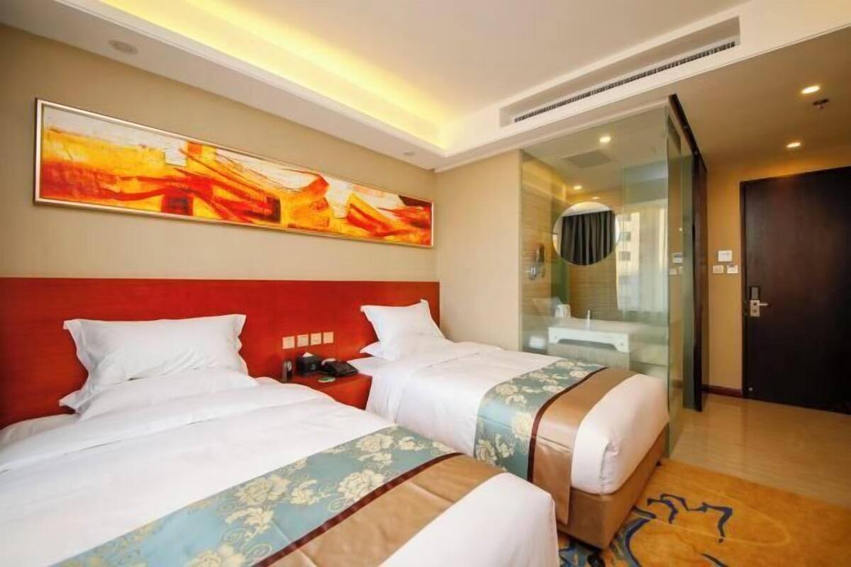 A hotel room with two beds, a red headboard, a large abstract painting, a glass partition, and a bathroom visible through the partition.