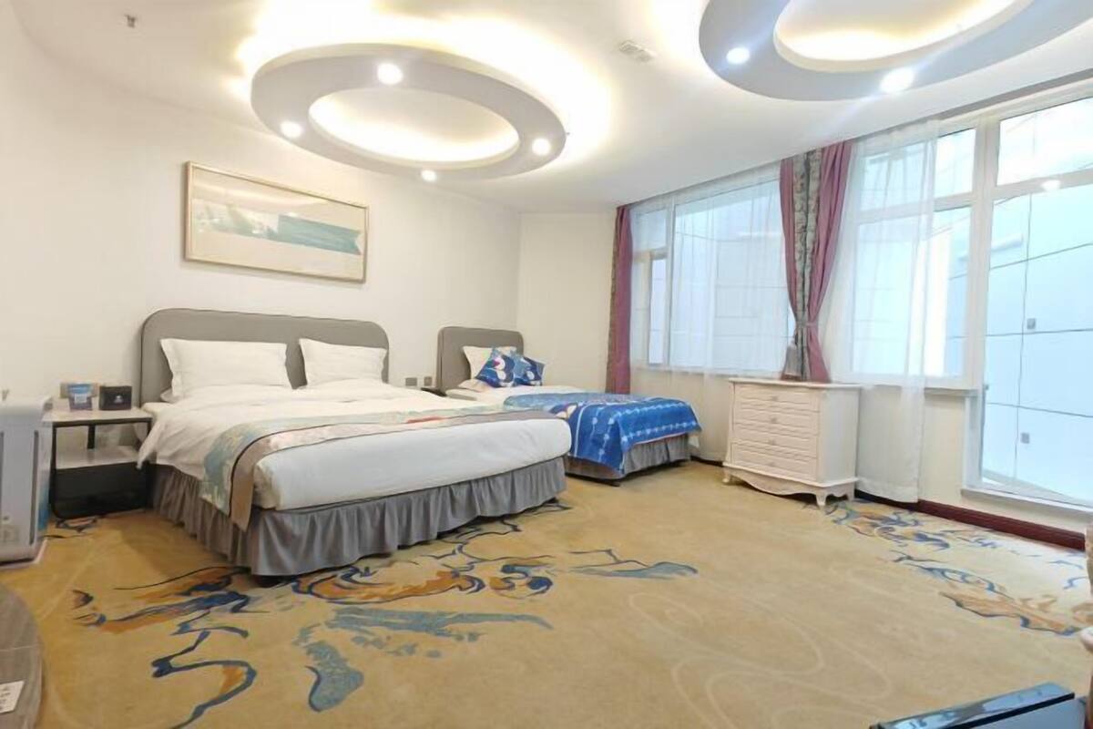 A hotel room with two beds, a large window with curtains, a carpet with a pattern, and a ceiling with circular light fixtures.