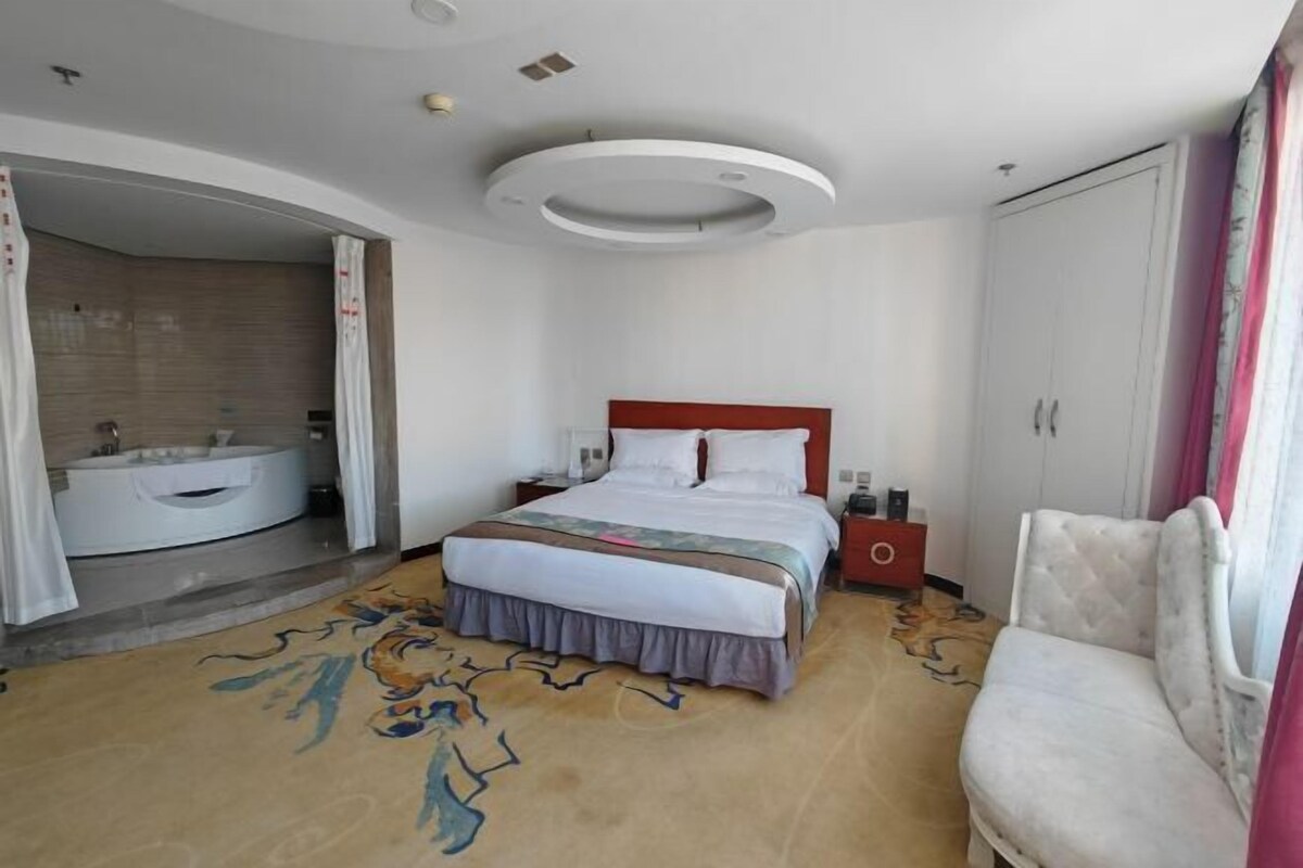 A hotel room with a bed, a vanity with a mirror, a red nightstand, a white wardrobe, and a chair.