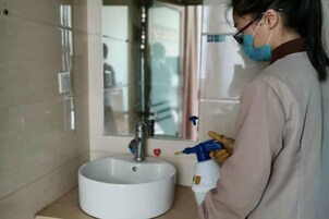Una persona en un baño, sosteniendo un atomizador, de pie cerca de un lavamanos con un espejo encima.