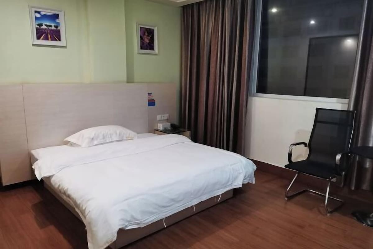 A hotel room with a bed, a chair, a nightstand, a window with curtains, and two wall-mounted pictures.