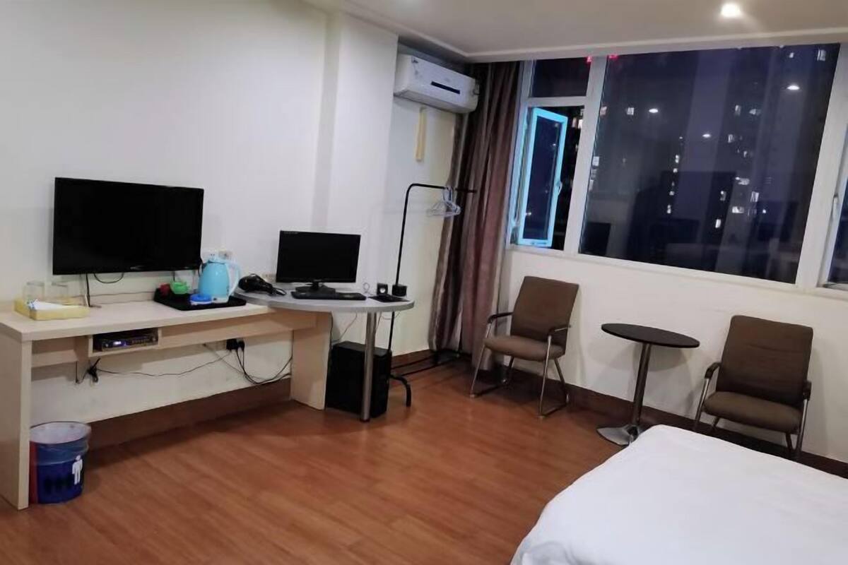 A hotel room with a bed, two TVs, a desk with a computer, a chair, a small table, and a window with a city view.