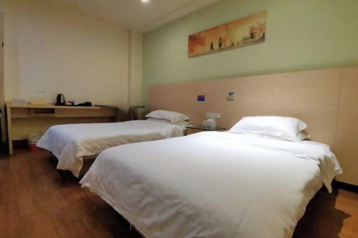 A hotel room with two single beds, a wooden headboard, a small table with a lamp, and a painting on the wall.