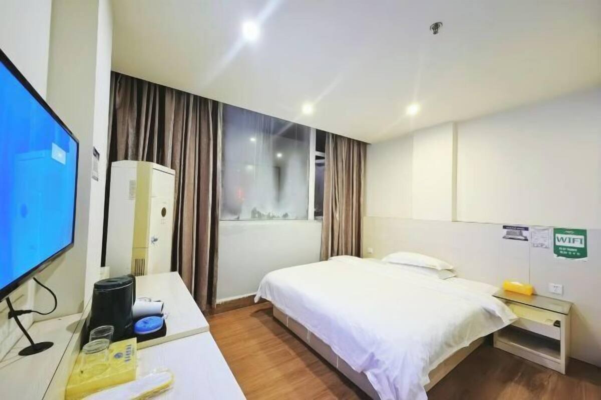 A hotel room with a bed, a desk with a computer, a window with curtains, and a sign indicating WiFi availability.