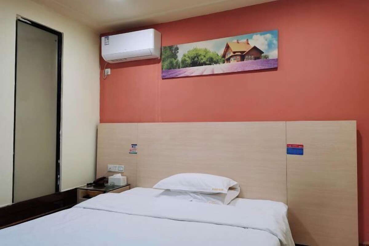 A hotel room with a bed, a nightstand, an air conditioning unit, and a painting on the wall.