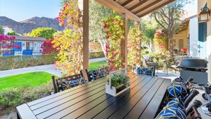 Um pátio externo com mesa e cadeiras de madeira, rodeado por plantas floridas e vista para montanhas.