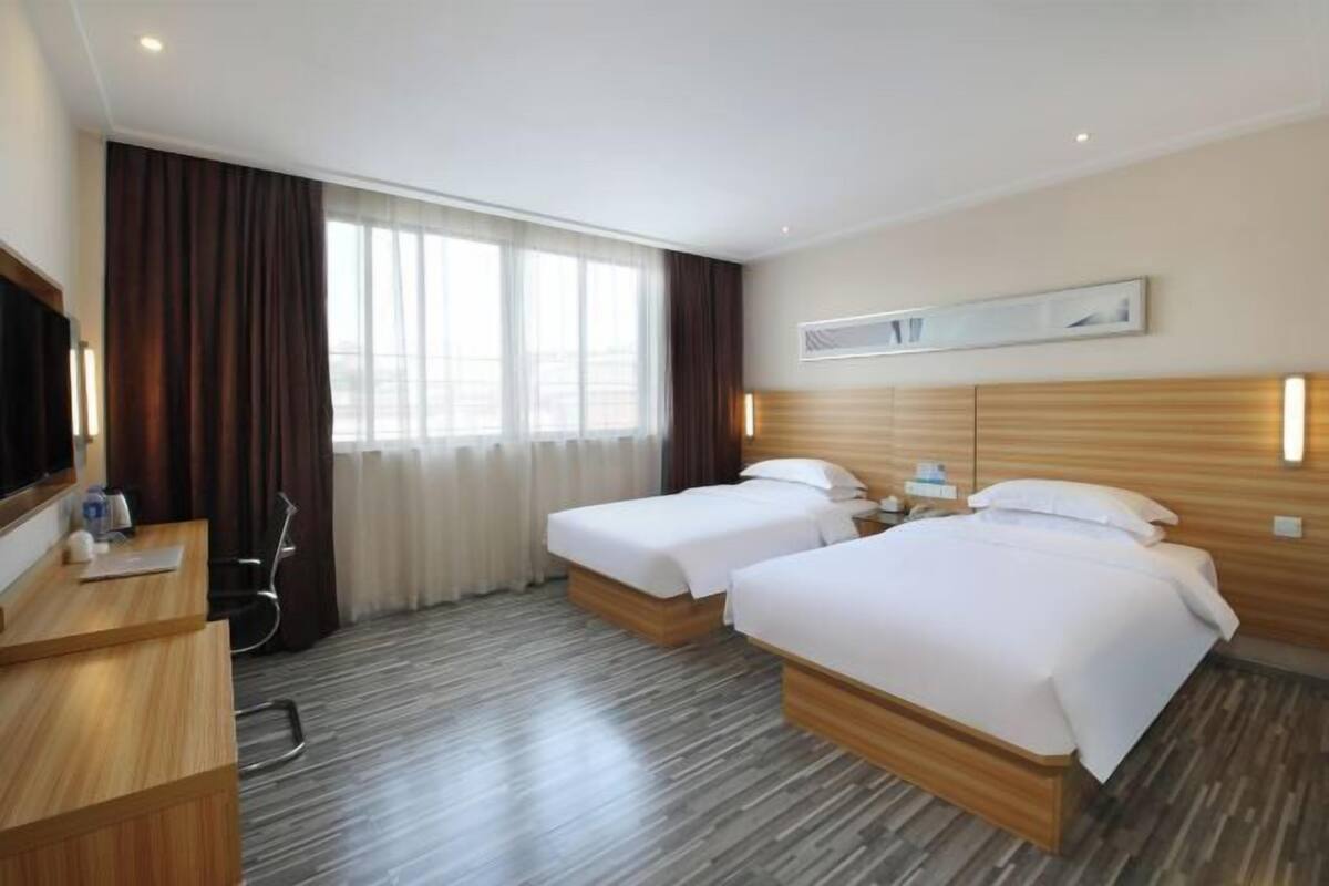 Ein Hotelzimmer mit zwei Betten, einem Schreibtisch, einem Fernseher und einem Fenster mit Vorhängen.