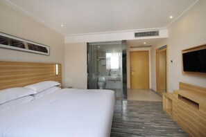 Ein Hotelzimmer mit einem großen Bett, einem hölzernen Kopfteil, einem an der Wand montierten Fernseher und einem durch eine Glastür einsehbaren Badezimmer.