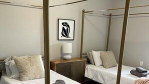 Una cama con dosel de madera y colcha blanca. Hay una mesita de noche con una lámpara y una toalla doblada. Una obra de arte enmarcada está en la pared sobre la cama.