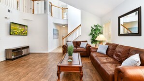 Een ruime woonkamer met een bruine leren bank, een houten salontafel en een tv op een standaard. Een trap met witte leuningen leidt naar een hoger gelegen verdieping.