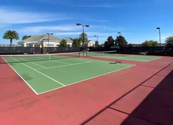 Una cancha de tenis con superficie roja y marcas verdes, rodeada por una valla y palmeras.