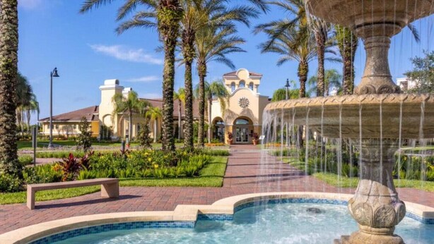 Una fuente en un área ajardinada con piscina, palmeras y un edificio con ventanas de arco en el fondo.
