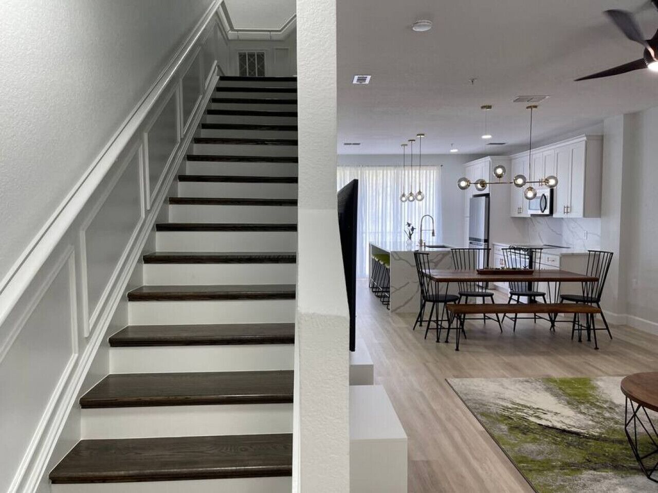 Un escalier avec des marches en bois foncé et des murs blancs, menant à un coin repas avec une table et des chaises en bois, et un ventilateur de plafond.
