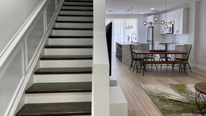 Una escalera con escalones de madera oscura y paredes blancas, que conduce a un comedor con una mesa y sillas de madera, y un ventilador de techo.