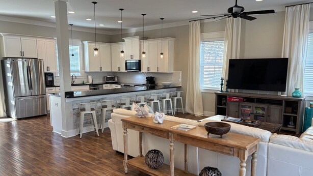 A modern kitchen with white cabinets, a stainless steel refrigerator, and a black countertop. There is a dining area with white chairs and a wooden table. A television is mounted on the wall, and a ceiling fan is visible.