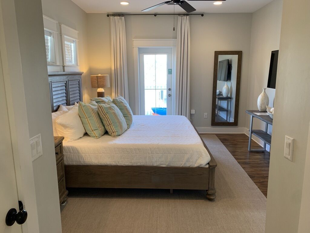 A bedroom with a large bed, a ceiling fan, a window with curtains, a bedside table with a lamp, and a mirror on the wall.