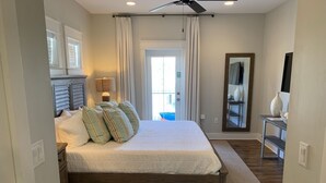 A bedroom with a large bed, a ceiling fan, a window with curtains, a bedside table with a lamp, and a mirror on the wall.