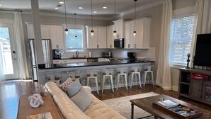 A modern kitchen with white cabinets, a stainless steel refrigerator, and a black countertop. There is a dining area with a beige sofa and a wooden coffee table.
