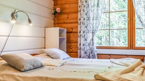 Acogedora habitación tipo cabaña de madera con cama, lámpara de noche, ventana con cortinas y un pequeño armario blanco.