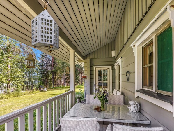 A covered porch with a glass table and wicker chairs, overlooking a grassy yard with trees.