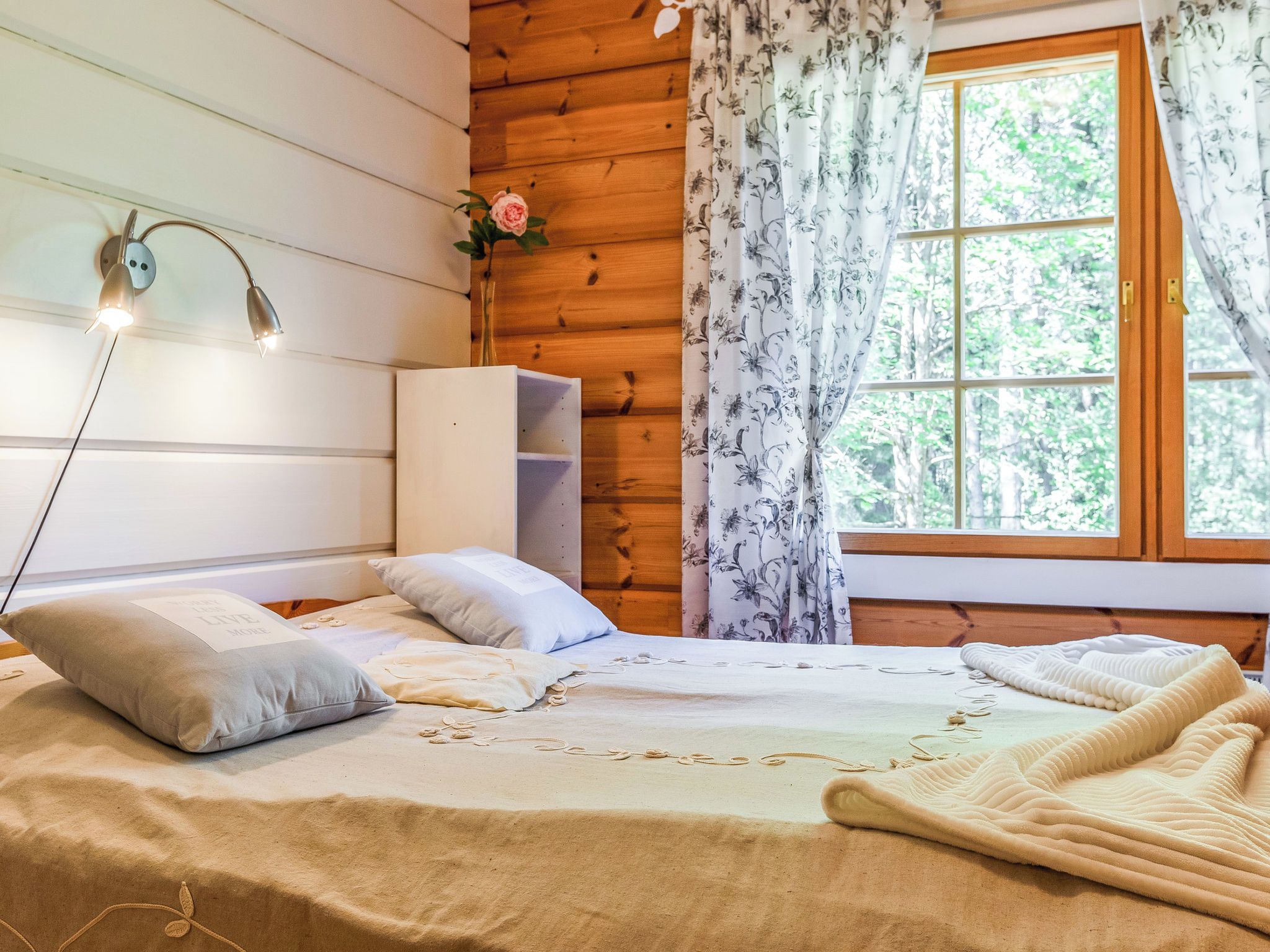 Une chambre à coucher dans une cabane en bois, avec un lit, des oreillers, une table de chevet, une lampe et une fenêtre avec des rideaux.