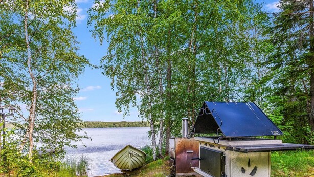 Una cabaña rústica con techo de metal, un bote pequeño y cimientos de concreto cerca de un lago rodeado de árboles.