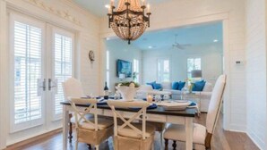 A dining area with a chandelier, white walls, and a view into a living room with a sofa and a TV. - WaterColor-Santa Rosa.  Golf Cart, 2 min from beach, Community Pools (Santa Rosa Beach)