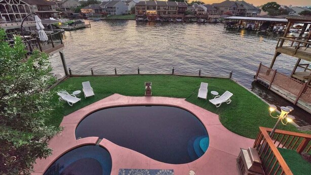 A swimming pool with a deck, surrounded by a grassy area and lounge chairs, overlooking a body of water with houses in the background. - Lakefront Retreat with Pool, Spa, Golf, Fishing,  Boat slip, Kayaks & Marina (Montgomery)