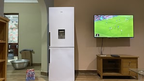 A kitchen with a white refrigerator, a wooden cabinet, a TV mounted on the wall, and a large window.