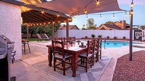 Un patio al aire libre con una mesa y sillas de madera, luces colgantes y una piscina al fondo.