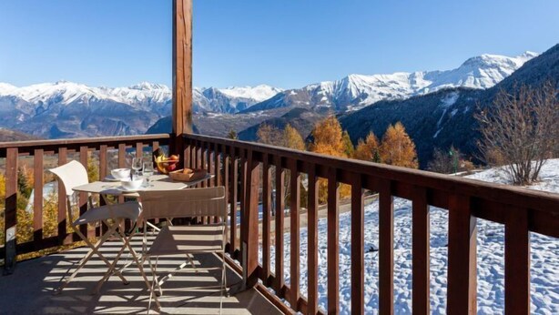 Eine Holzterrasse mit Tisch und Stühlen mit Blick auf schneebedeckte Berge und Herbstbäume.