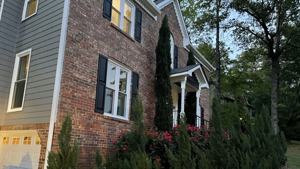 A two-story house with a brick facade, white trim, and black shutters. There is a covered porch with a railing and a small garden with red flowers and green shrubs. - Welcome Home!! (Marietta)