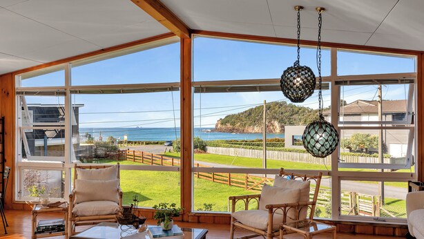 Quarto amplo com grandes janelas proporcionando vista para o mar e um deck de madeira. Há cadeiras de vime e uma mesa de centro com objetos decorativos.