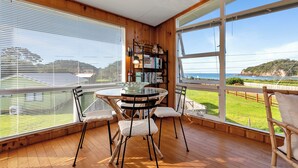 Um deck de madeira com uma mesa redonda e cadeiras, uma estante e vista para o mar.