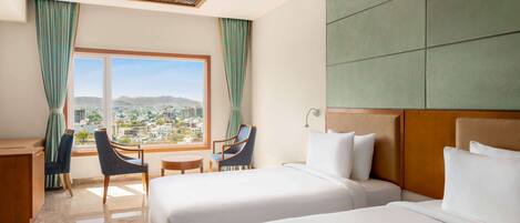 A hotel room with two beds, a desk, a chair, and a view of a cityscape through a large window.