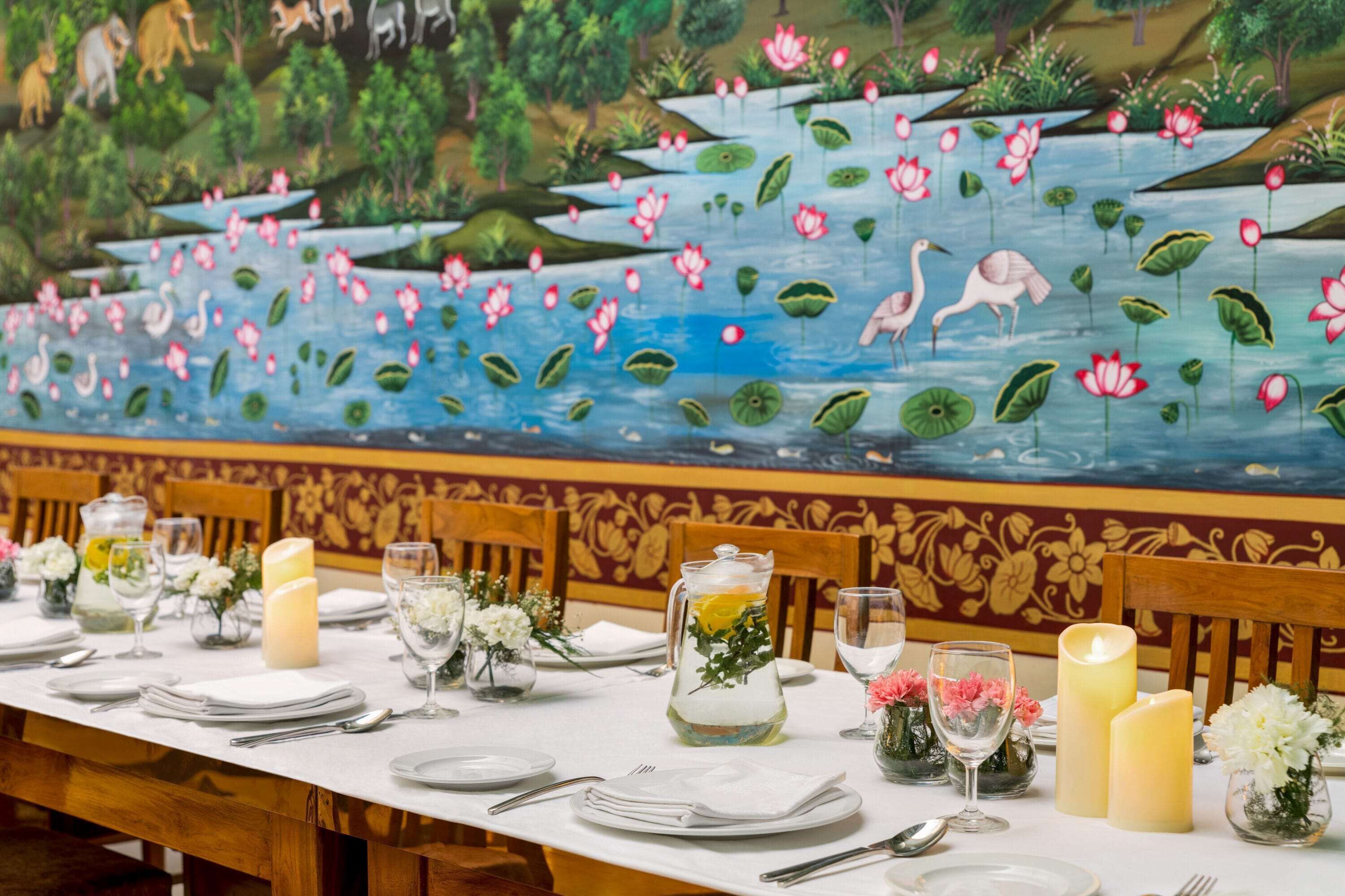 A dining table set with white plates, silverware, and candles, with a mural of a pond and animals in the background.
