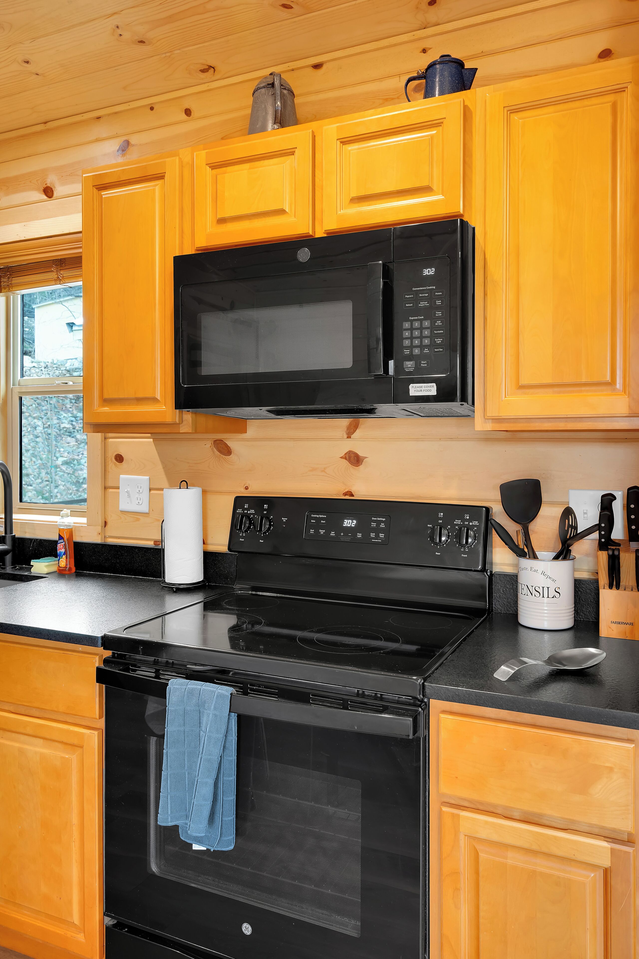 Une cuisine avec des meubles en bois, un micro-ondes noir, une cuisinière et une fenêtre donnant sur les arbres.